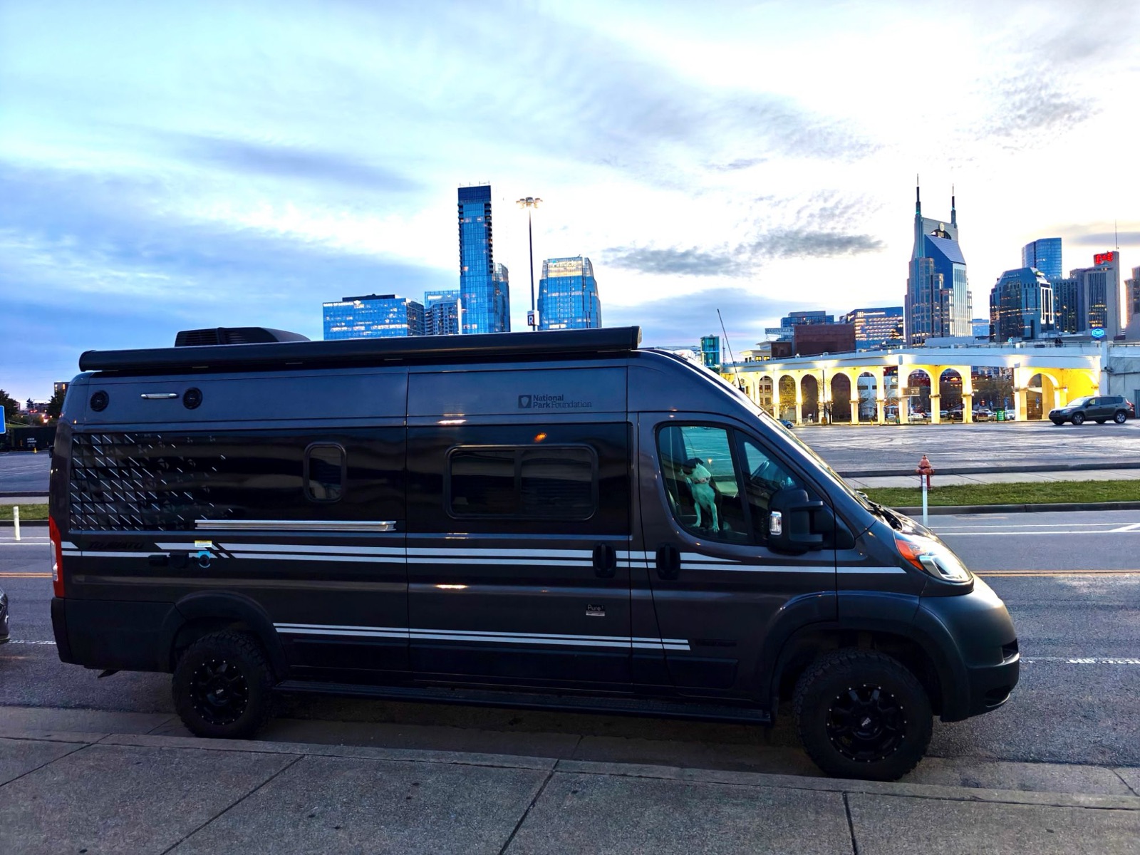 Stanley, a black 2020 Winnebago Travato 59GL National Park Edition, parked on a city street at dusk with a skyline behind it and a dog looking out the driver's window.